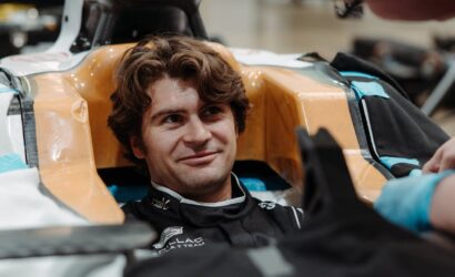 Smiling man sits in a race car cockpit with orange padded sides, wearing a racing suit and helmet off nearby.