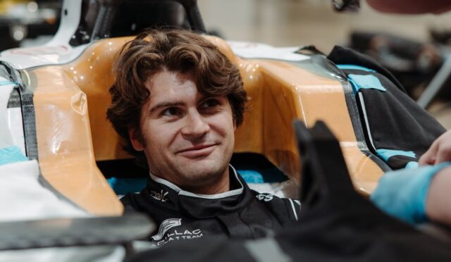 Smiling man sits in a race car cockpit with orange padded sides, wearing a racing suit and helmet off nearby.