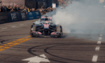 Pink and blue Formula 1 car performs a burnout on a race track, smoke billowing behind as crowds watch from the stands.