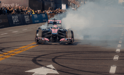 Pink and blue Formula 1 car performs a burnout on a race track, smoke billowing behind as crowds watch from the stands.