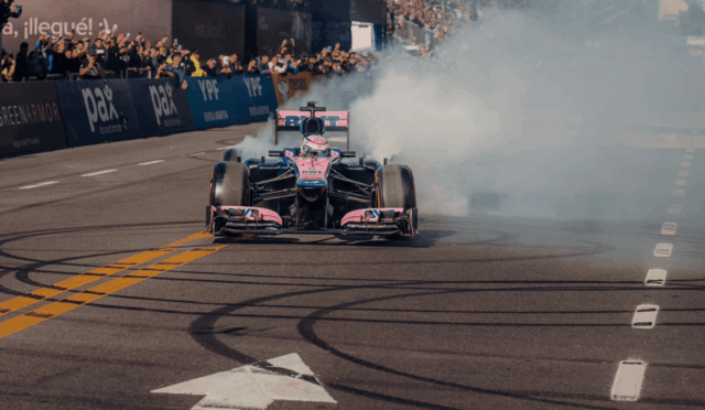 Pink and blue Formula 1 car performs a burnout on a race track, smoke billowing behind as crowds watch from the stands.