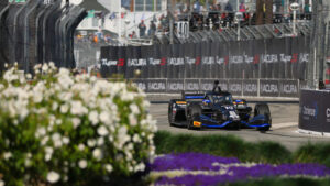 Blue and black Formula race car speeding along a track with barrier walls, colorful flowers in the foreground, and spectators in the stands in the background.