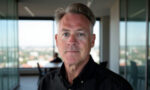 Portrait of a middle-aged man with gray hair in a black shirt, indoors with a city skyline behind him in a glass-walled office setting.