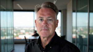 Portrait of a middle-aged man with gray hair in a black shirt, indoors with a city skyline behind him in a glass-walled office setting.
