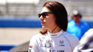 Female race car driver in a white racing suit and dark sunglasses stands on the pit lane with a blurred race track in the background.