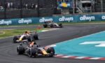 Formula 1 cars race around a bend on a circuit, leading car in front with others close behind and sponsor boards along the barrier.