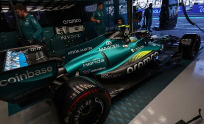 Teal Formula 1 car in the pit with crew members and sponsor logos like Aramco, Honda, and Aston Martin visible nearby.