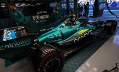 Teal Aston Martin Formula 1 car in the pit garage with engineers in teal uniforms nearby and equipment around.