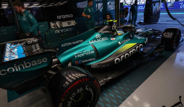 Teal Aston Martin Formula 1 car in the pit garage with engineers in teal uniforms nearby and equipment around.