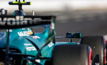 Close-up of a teal Formula 1 car on the track, showing the rear wing, cockpit area, and blurred background indicating speed.