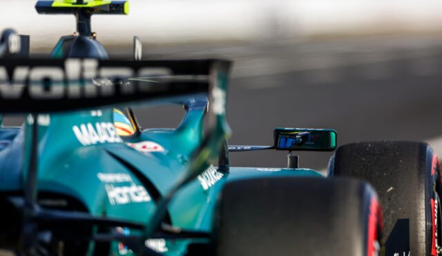 Close-up of a teal Formula 1 car on the track, showing the rear wing, cockpit area, and blurred background indicating speed.