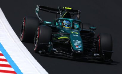 teal Formula 1 car on a racetrack, leaning into a turn with sponsor logos visible on the bodywork and tires in motion