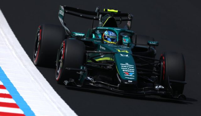 teal Formula 1 car on a racetrack, leaning into a turn with sponsor logos visible on the bodywork and tires in motion