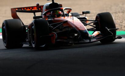 Red-orange Formula 1 car cornering on track, low angle shot emphasizing front wing and tire grip on tarmac curve.