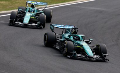 Two teal Formula 1 cars race side by side on a curving track, negotiating a bend with grass along the edge.