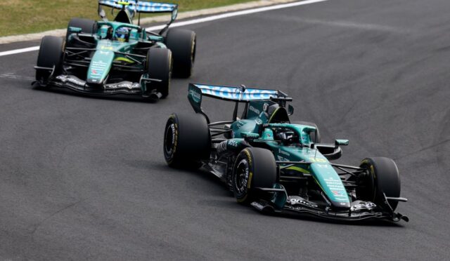 Two teal Formula 1 cars race side by side on a curving track, negotiating a bend with grass along the edge.