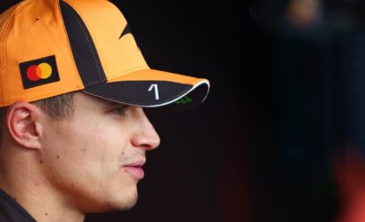 Profile view of a man wearing an orange cap with a Mastercard logo, looking to the right against a dark background prep area.