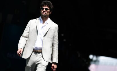 Man in a light gray suit and white shirt wearing sunglasses, standing on a dark stage.