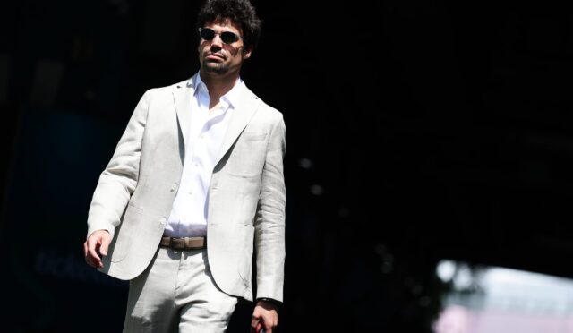 Man in a light gray suit and white shirt wearing sunglasses, standing on a dark stage.
