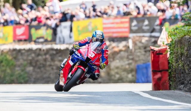 Motorcycle racer leaning into a corner on a road race, bike number 3 amid cheering crowds and barriers in the background.