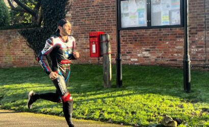 Male jogger in a racing suit runs past a brick wall, red post box, and parish council noticeboard labeled 'OSBASTON PARISH COUNCIL'.