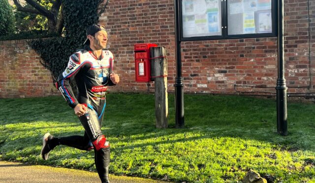 Male jogger in a racing suit runs past a brick wall, red post box, and parish council noticeboard labeled 'OSBASTON PARISH COUNCIL'.