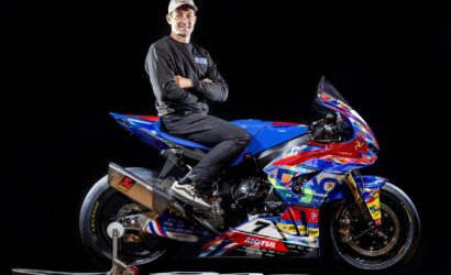 Professional motorcycle racer posing with a vibrant blue, red, and white race bike against a black studio background, arms crossed.