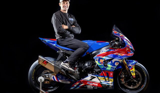Professional motorcycle racer posing with a vibrant blue, red, and white race bike against a black studio background, arms crossed.