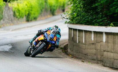 Moto racer leaning into a sharp turn on a racing bike with colorful livery, helmet visor visible on a narrow outdoor road edge wall beside greenery.