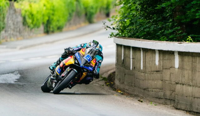 Moto racer leaning into a sharp turn on a racing bike with colorful livery, helmet visor visible on a narrow outdoor road edge wall beside greenery.