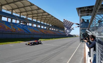 Formula 1 car crosses a track near the finish line as a checkered flag is waved by officials in a pit box while photographers capture the moment under a blue sky.