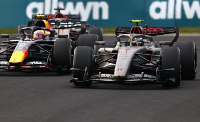 Three Formula 1 cars race side by side on a track, with a silver Mercedes in the foreground and Red Bull and others behind.