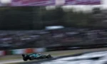 Open-wheel race car speeding along a blurred race track with grandstands in the background, teal color visible on the car.