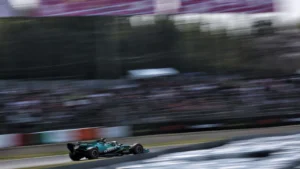 Open-wheel race car speeding along a blurred race track with grandstands in the background, teal color visible on the car.