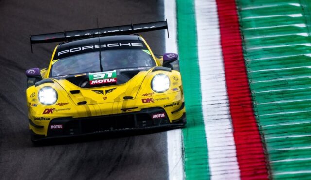 Yellow Porsche race car on a track with green, white, and red curbs, headlights on as it corners.