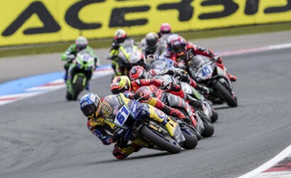 Group of motorcycle racers leaning into a sharp corner on a track, colorful helmets and suits, blurred yellow advertising boards in the background.
