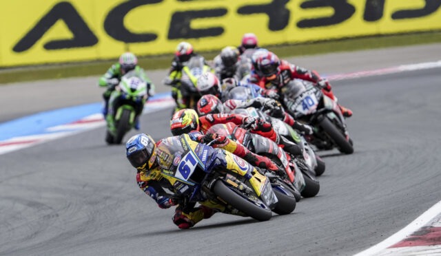 Group of motorcycle racers leaning into a sharp corner on a track, colorful helmets and suits, blurred yellow advertising boards in the background.