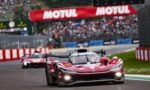 Red prototype race car leading a moment on a racetrack with a Motul banner and blurred cheering crowd in the background.
