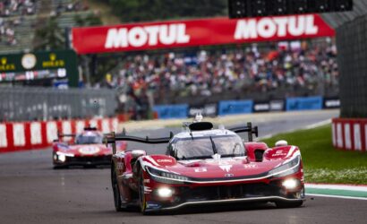 Red prototype race car leading a moment on a racetrack with a Motul banner and blurred cheering crowd in the background.