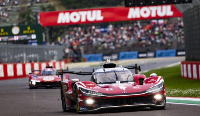Red prototype race car leading a moment on a racetrack with a Motul banner and blurred cheering crowd in the background.
