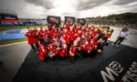 Motorsport team in red uniforms posing for a group photo on the race track with sponsor banners in the background.