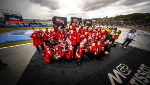 Motorsport team in red uniforms posing for a group photo on the race track with sponsor banners in the background.