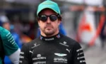 Male race car driver in a black sponsor-laden suit and teal cap, wearing sunglasses, facing the camera in a blurred pit area.