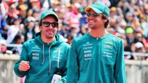 Two men in teal team jackets with sponsor logos walk side by side at a crowded event, wearing caps and sunglasses.
