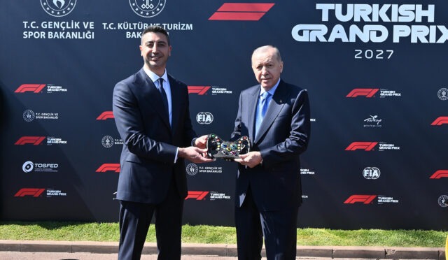 Two men in suits exchange a decorative trophy in front of a Turkish Grand Prix 2027 backdrop, smiling at the camera вЂ” formal award ceremony scene.