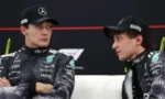 Two Mercedes F1 drivers in black racing suits and caps sit together, talking during an interview on a podium backdrop.