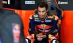 MotoGP rider in a Red Bull KTM jumpsuit sits in a team chair, hands clasped, focused before a race in the pit area.