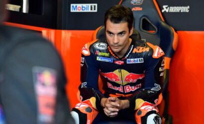 MotoGP rider in a Red Bull KTM jumpsuit sits in a team chair, hands clasped, focused before a race in the pit area.