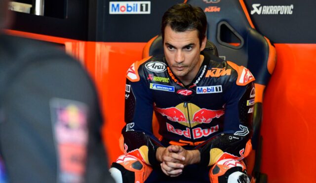 MotoGP rider in a Red Bull KTM jumpsuit sits in a team chair, hands clasped, focused before a race in the pit area.