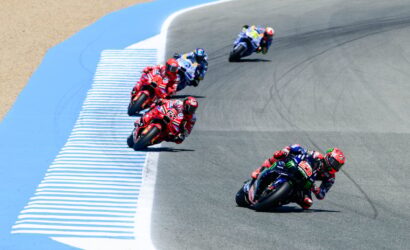 Racing motorcycles lean into a curve on a race track, multiple riders in colorful gear leading the pack.